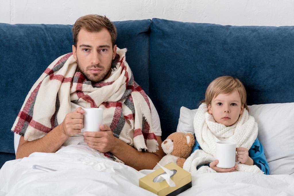 padre malato e figlio in sciarpe con tazze di bevanda calda seduto a letto e guardando la fotocamera
 - Foto, immagini