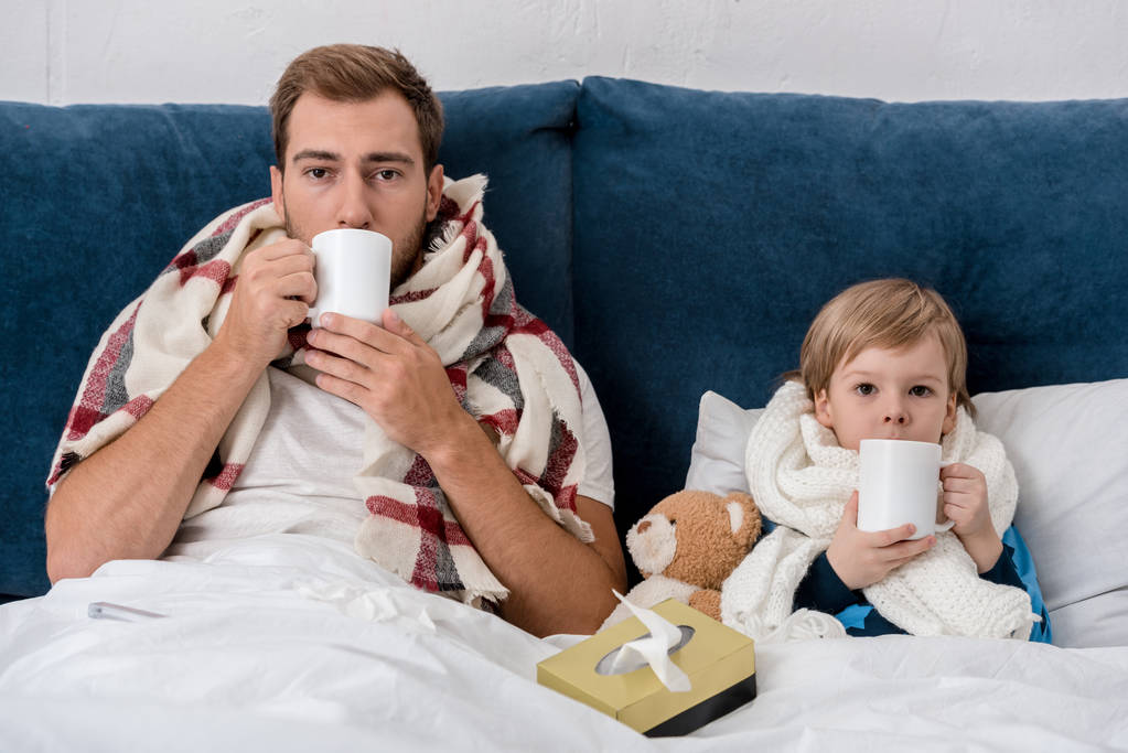 sairas isä ja poika huiveissa juo teetä, kun istuu sängyssä ja katselee kameraa
 - Valokuva, kuva