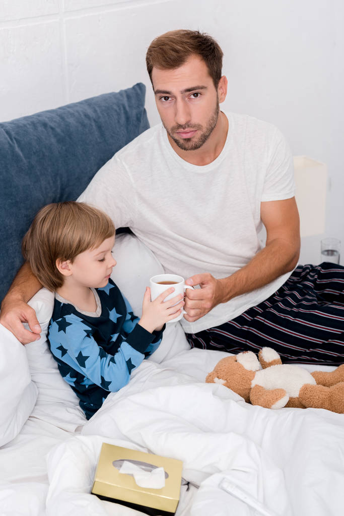 young father giving cup of hot drink to his son in bed and looking at camera - Photo, Image