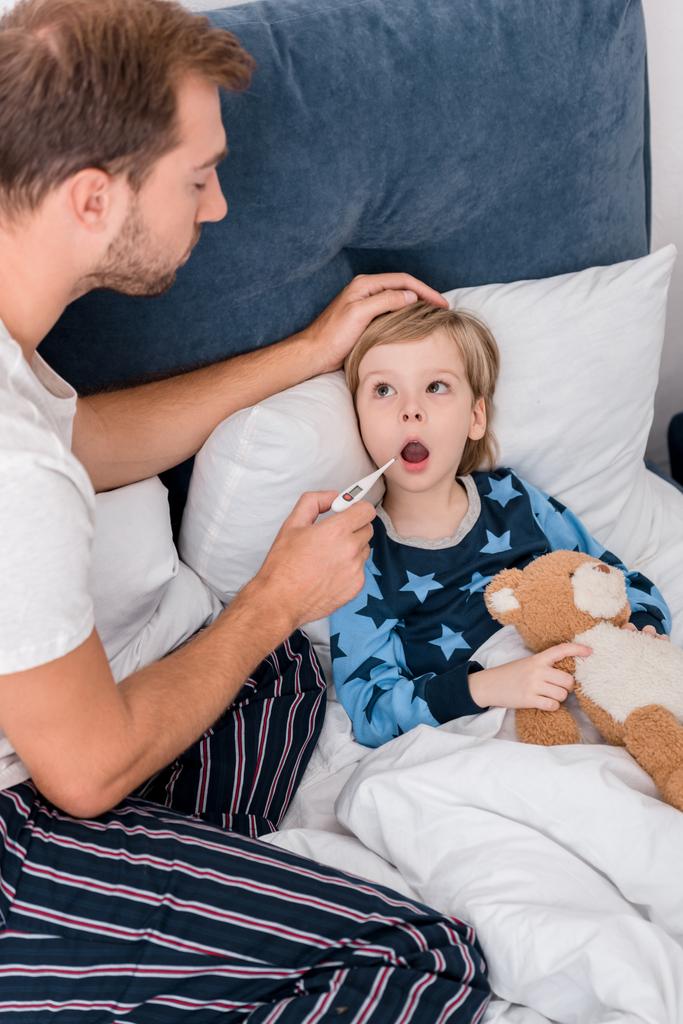 alto ángulo vista de padre comprobar oral temperatura de hijo mientras él acostado en cama
 - Foto, imagen