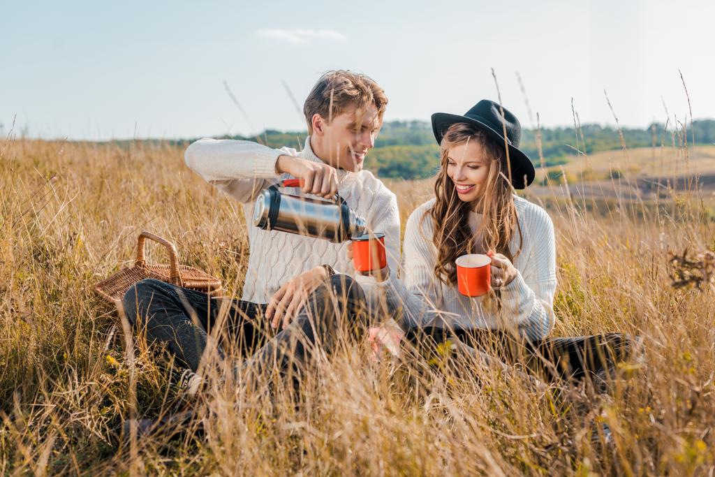 happy boyfriend pouring hot drink from thermos into cups for girlfriend on rural meadow - Photo, Image