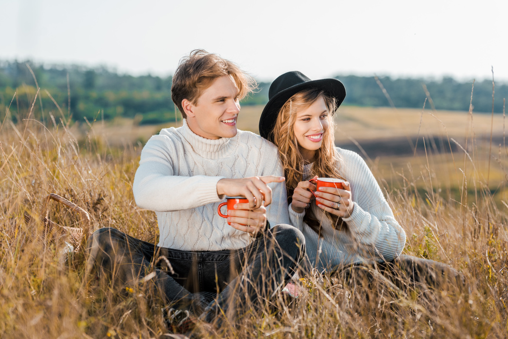 sonriente pareja con bebidas calientes mirando hacia el prado rural
 - Foto, Imagen