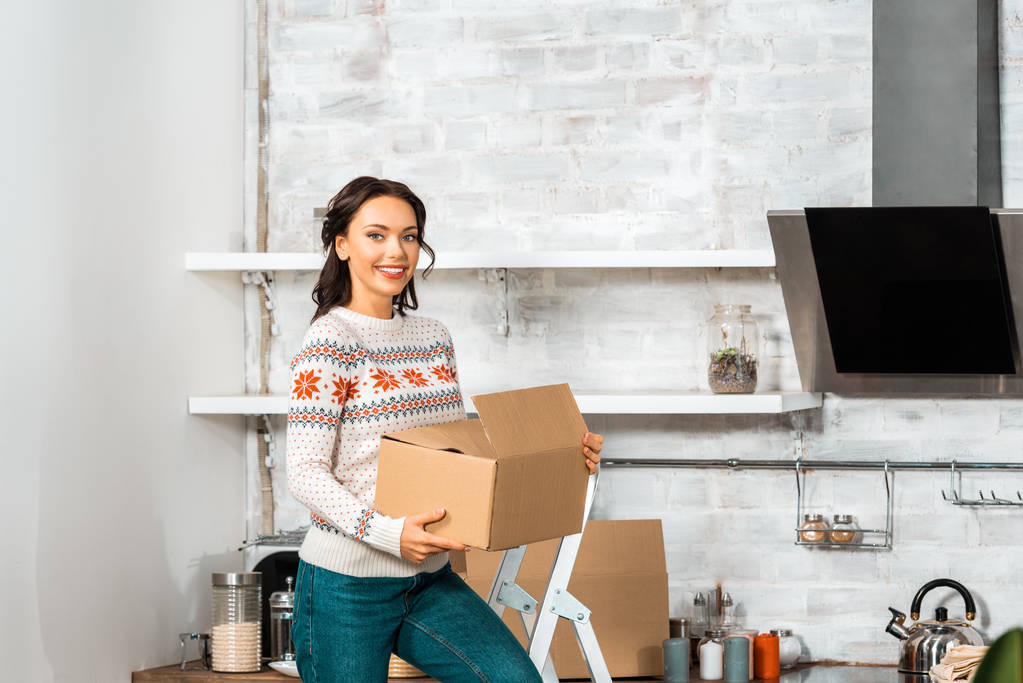 ευτυχισμένη γυναίκα στέκεται πάνω σε Σκάλα, κουτί από χαρτόνι στην κουζίνα στο νέο σπίτι  - Φωτογραφία, εικόνα
