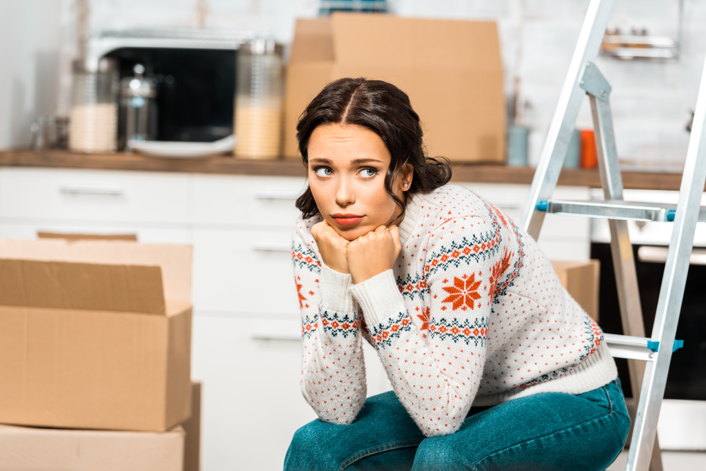 сумна красива жінка сидить на драбині на кухні з картонними коробками під час переїзду в новому будинку
 - Фото, зображення