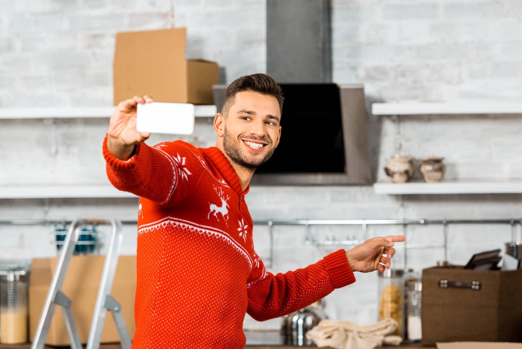 笑みを浮かべて男のスマート フォンと新しい家に移転中に段ボール箱が付いている台所で指で指す selfie を取って  - 写真・画像