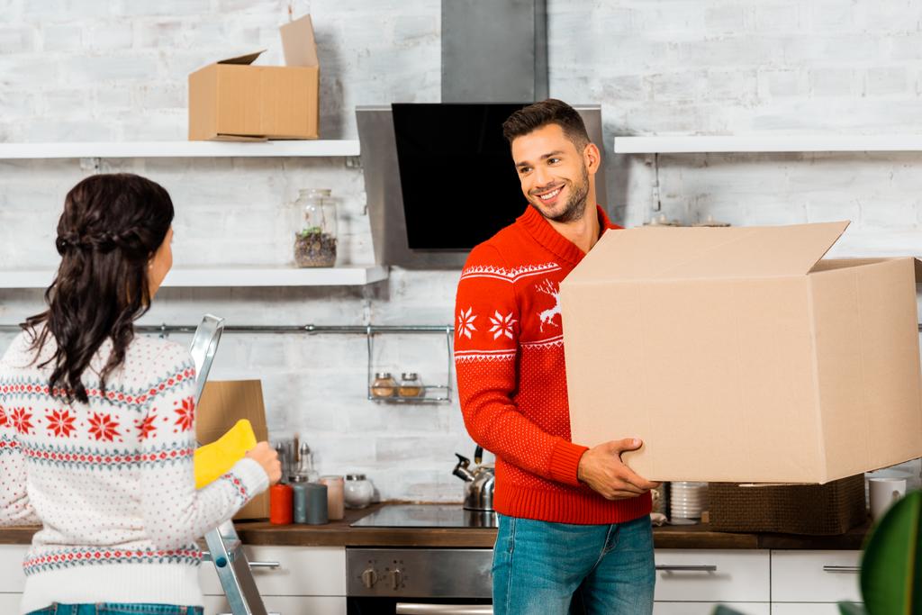 веселый мужчина держит картонную коробку и разговаривает с подругой на кухне в новом доме
  - Фото, изображение
