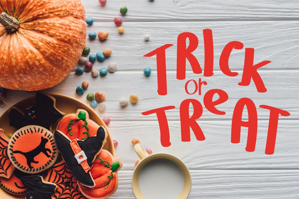 top view of pumpkin, plate with halloween cookies, candies and cup with milk on wooden table  with "trick or treat" lettering - Photo, Image