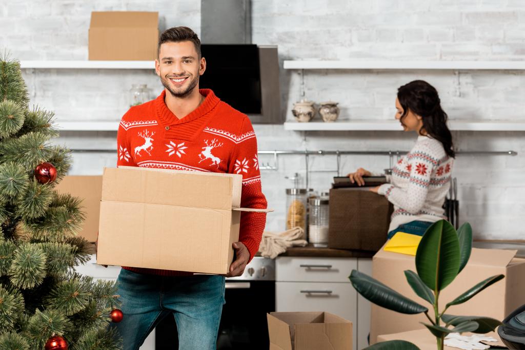 自宅の台所で後ろに立っている彼のガール フレンド、クリスマス ツリーを飾るためのつまらないものと段ボール箱を抱きかかえた - 写真・画像