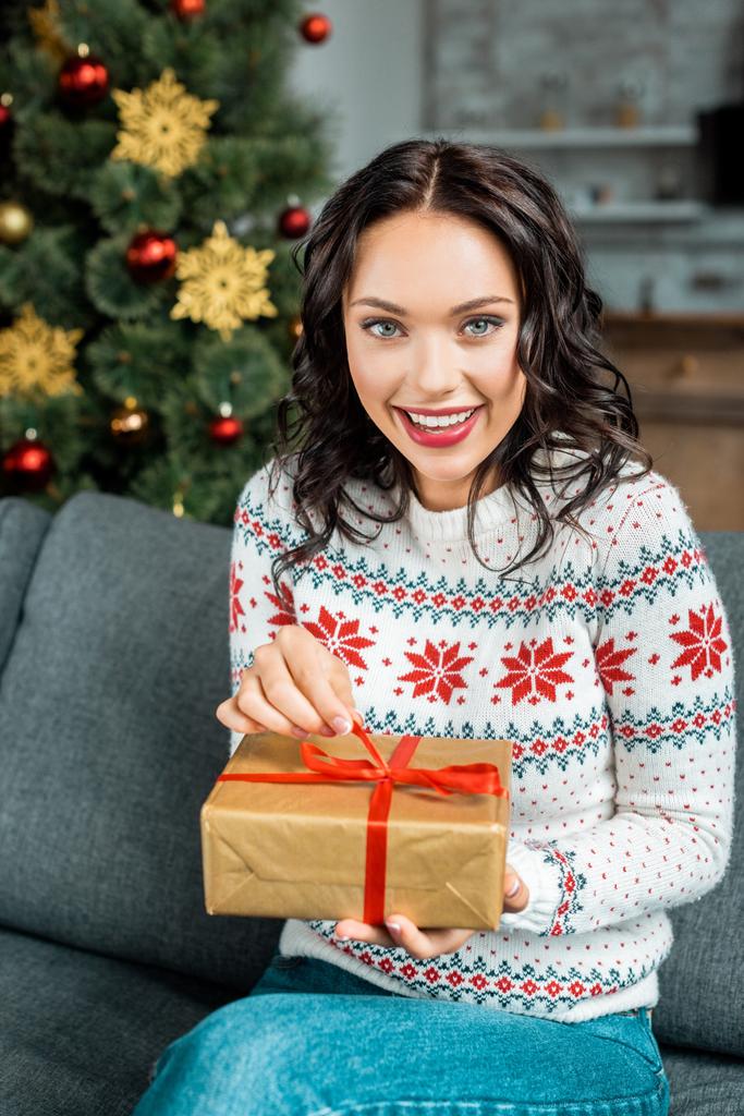mujer joven sonriente con caja de regalo en el sofá cerca del árbol de Navidad en casa
 - Foto, Imagen
