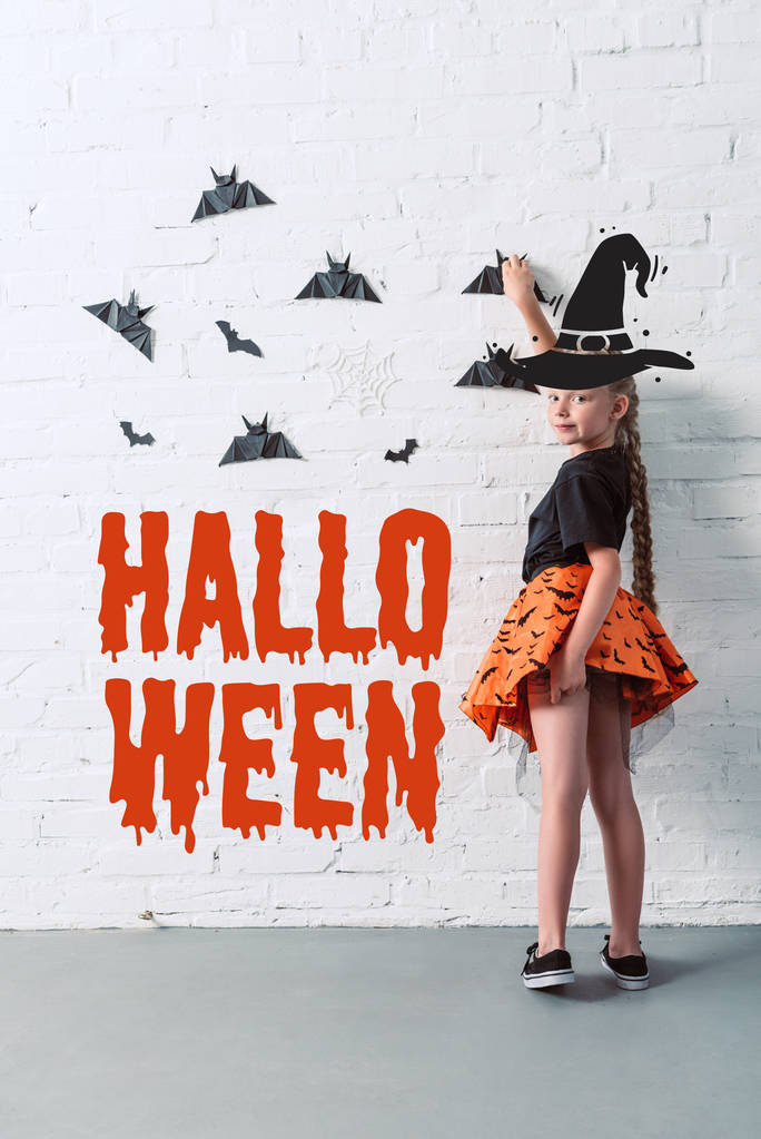 vue arrière de l'enfant en jupe suspendue chauves-souris en papier noir sur un mur de briques blanches, avec lettrage "halloween"
 - Photo, image