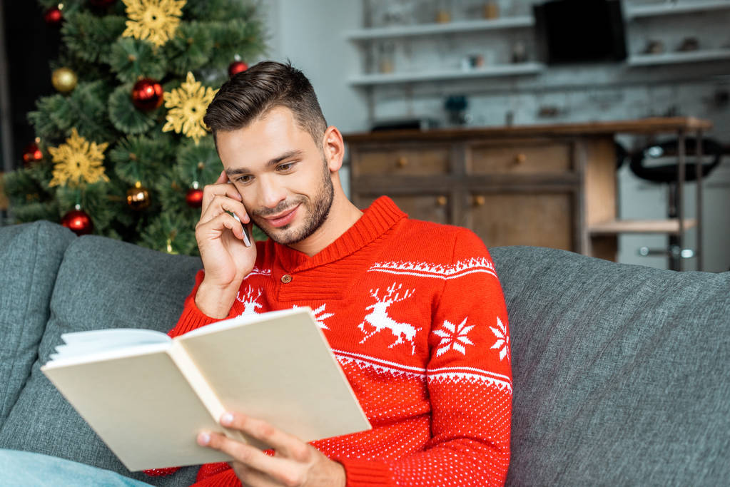 όμορφος νεαρός άνδρας, διαβάζοντας το βιβλίο και να μιλάμε για το smartphone κοντά στο χριστουγεννιάτικο δέντρο στο σπίτι  - Φωτογραφία, εικόνα