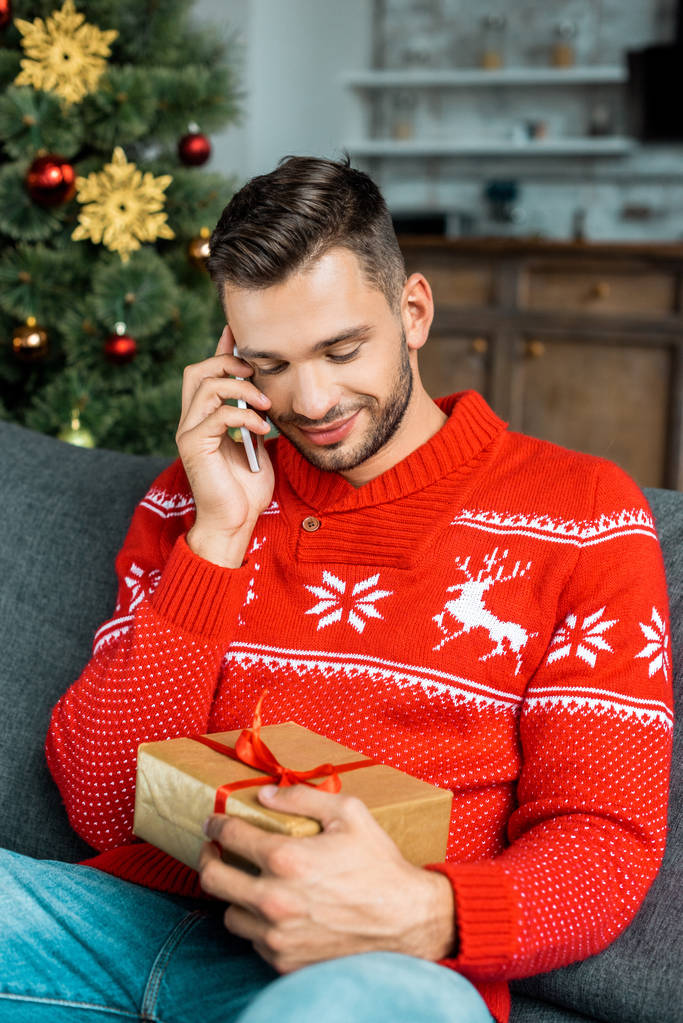 улыбающийся мужчина разговаривает по смартфону и держит подарочную коробку на диване возле рождественской елки дома
  - Фото, изображение