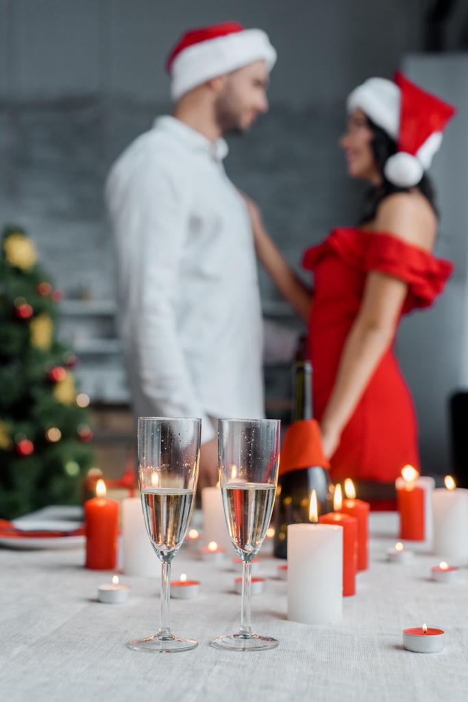 foco seletivo de copos de champanhe na mesa servida com velas e casal em chapéus de Natal em pé atrás em casa
 - Foto, Imagem