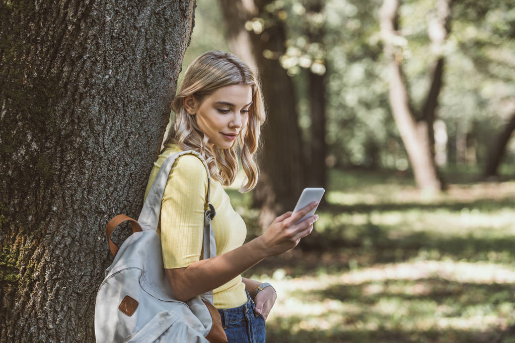 young woman with backpack on shoulder using smartphone in forest - Photo, Image