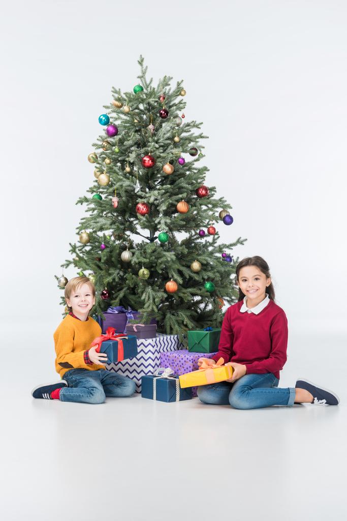 Geschwister sitzen mit Geschenken am Weihnachtsbaum und schauen vereinzelt in die Kamera - Foto, Bild