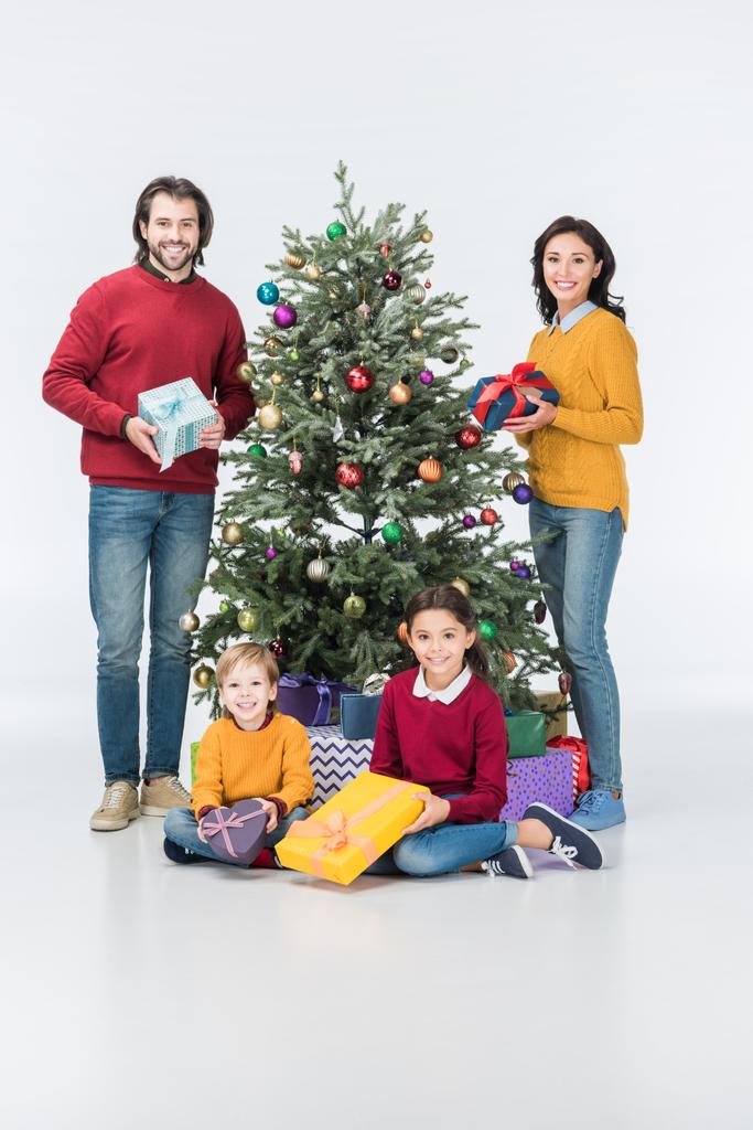 Genitori felici con scatole regalo in piedi mentre fratelli seduti vicino all'albero di Natale isolato su bianco
 - Foto, immagini