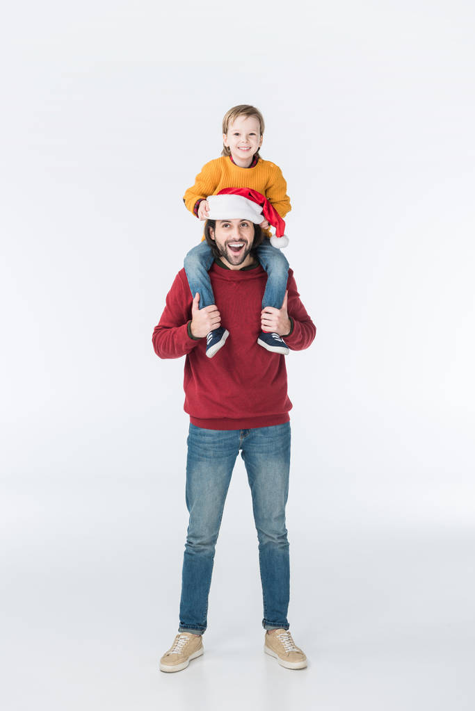 Gelukkig vader in Kerstman hoed zoon dragen op schouders geïsoleerd op wit - Foto, afbeelding