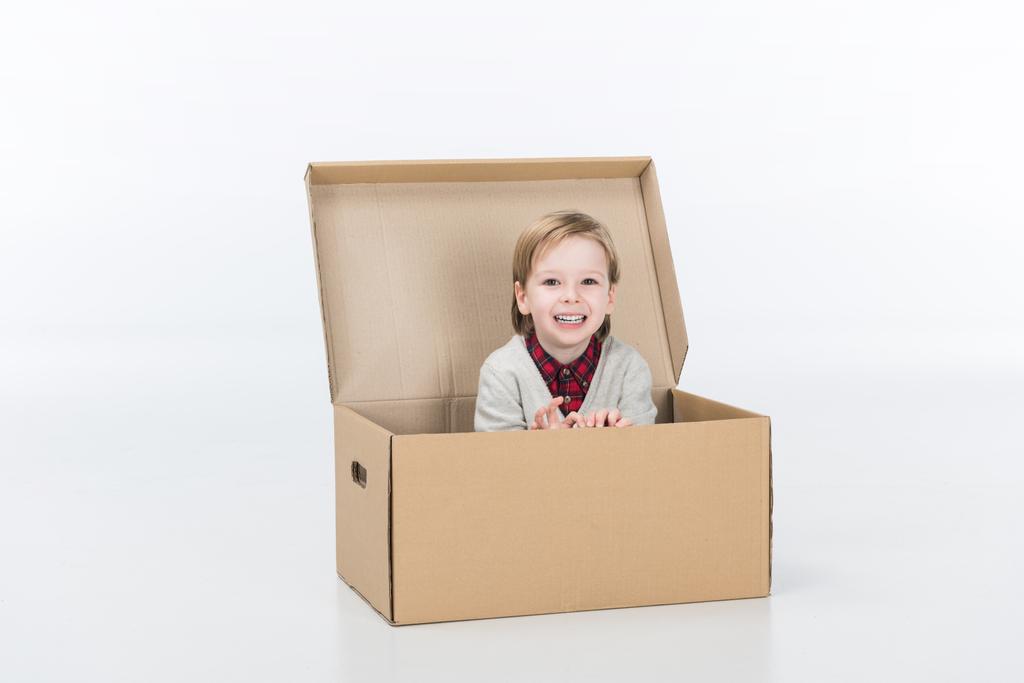 χαμογελαστό αγόρι καθιστός σε κουτί από χαρτόνι που απομονώνονται σε λευκό - Φωτογραφία, εικόνα
