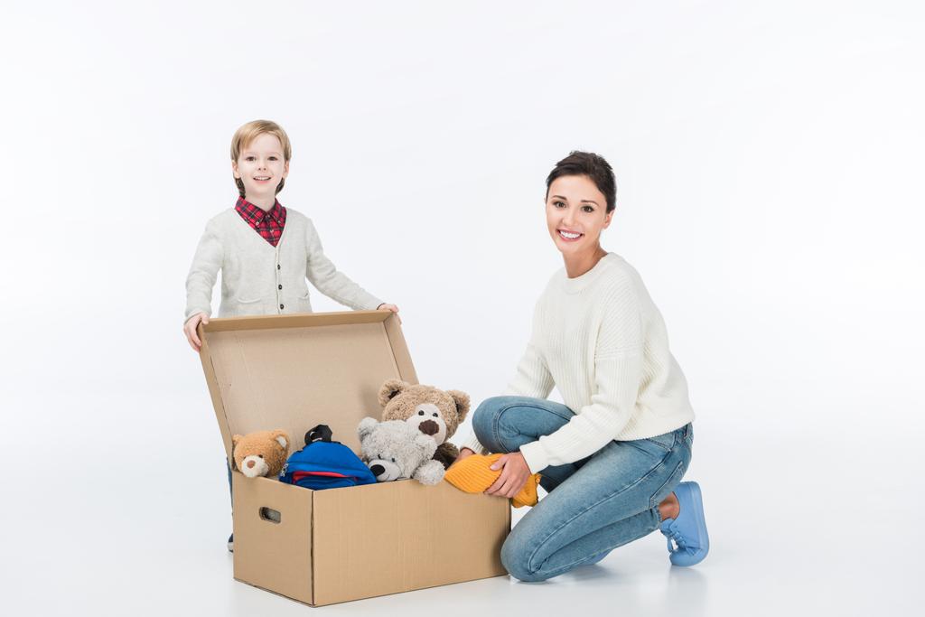 mãe sorridente com filho sentado perto de caixa de papelão com brinquedos e olhando para a câmera isolada no branco
  - Foto, Imagem