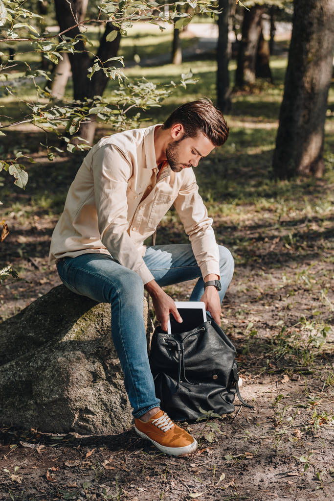 νεαρός άνδρας βάζοντας ψηφιακή δισκίο σε σακίδιο στο πάρκο - Φωτογραφία, εικόνα