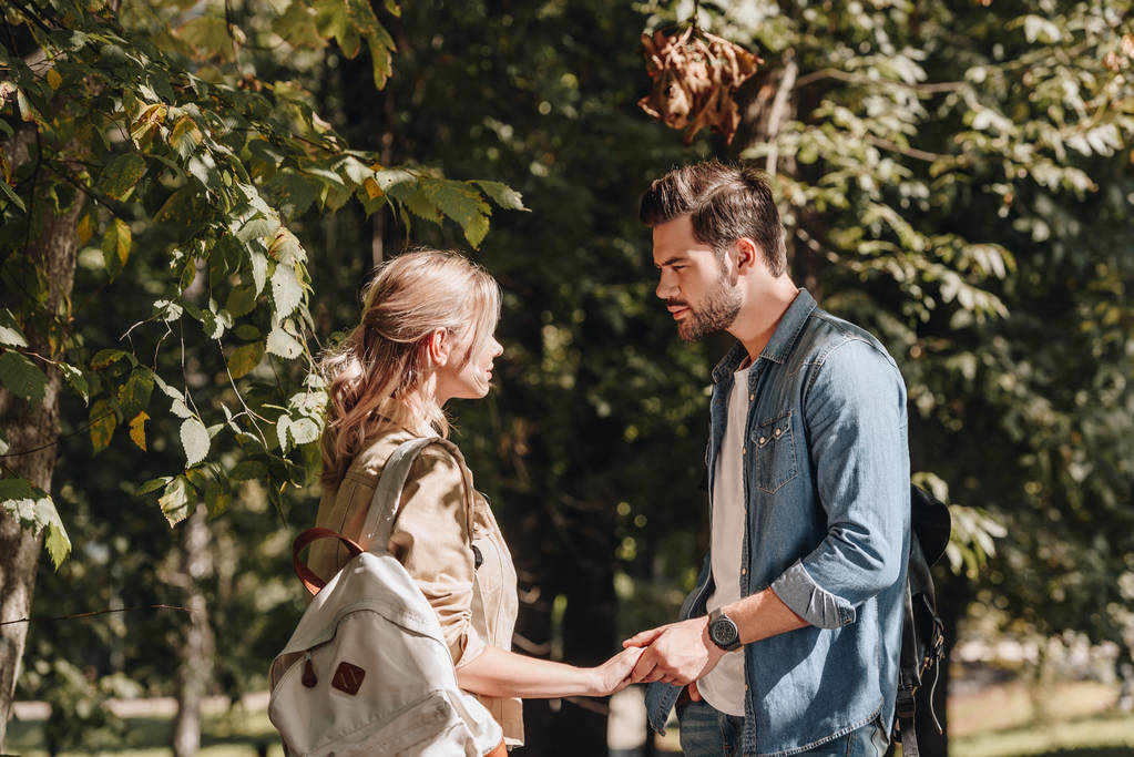 vista lateral de pareja joven con mochilas cogidas de la mano y mirándose en el parque
 - Foto, Imagen