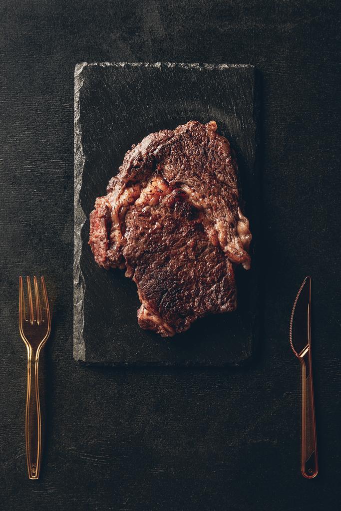 kızarmış biftek üstten görünüm siyah ahşap tahta, plastik bıçak ve çatal mutfakta masada - Fotoğraf, Görsel