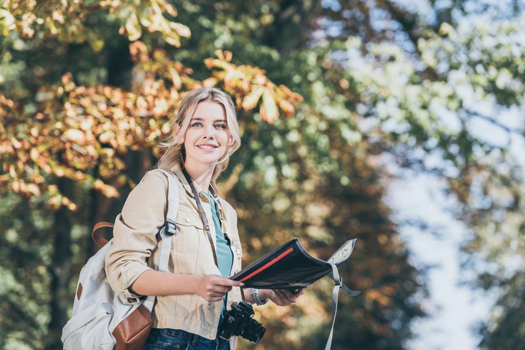 портрет молодого улыбающегося путешественника с рюкзаком, фотокамерой и картой в парке
 - Фото, изображение
