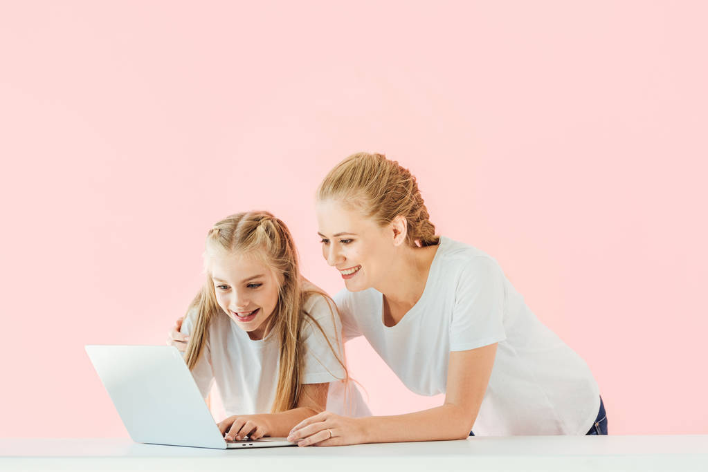 χαμογελαστός, μητέρα και κόρη στην λευκά μπλουζάκια χρησιμοποιώντας φορητό υπολογιστή μαζί απομονώνονται σε ροζ - Φωτογραφία, εικόνα