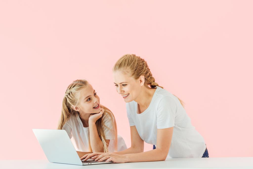 mooie moeder en dochter in witte t-shirts met behulp van laptop samen geïsoleerd op roze - Foto, afbeelding