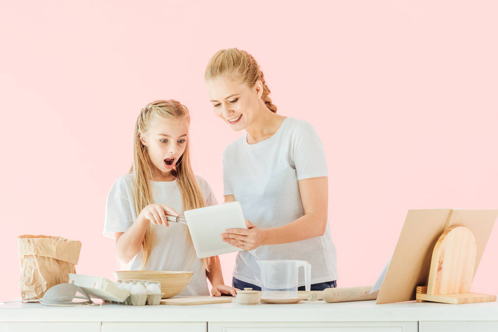 Χαμογελώντας η μητέρα και έκπληκτος κόρη στα λευκά μπλουζάκια χρησιμοποιώντας tablet ενώ μαγείρεμα απομονώνονται σε ροζ - Φωτογραφία, εικόνα