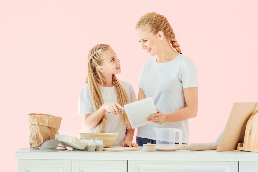 happy young mother and daughter in white t-shirts using tablet and looking at each other while cooking isolated on pink - Photo, Image