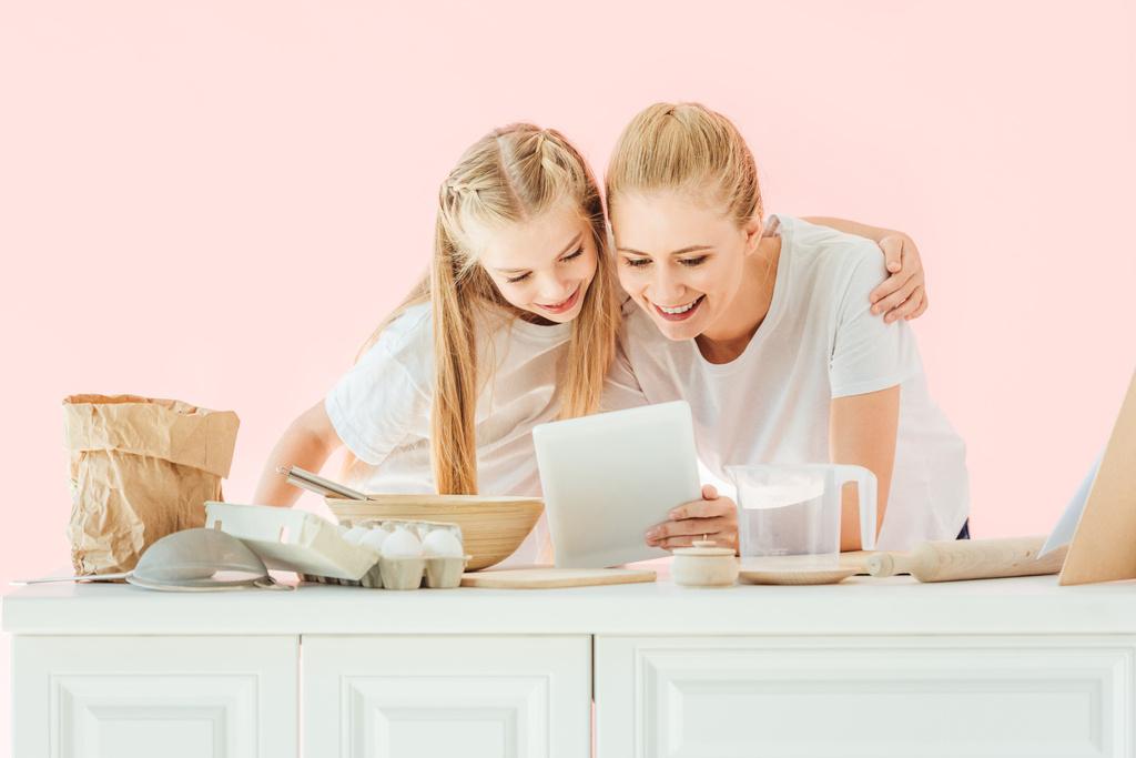 lachende Mutter und Tochter in weißen T-Shirts mit Tablet beim Kochen isoliert auf rosa - Foto, Bild