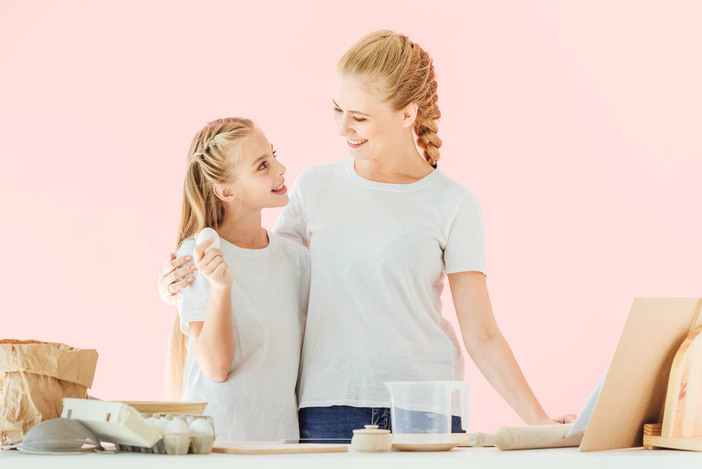 молодая мать и очаровательная маленькая дочь в белых футболках, глядя друг на друга во время приготовления пищи изолированы на розовый
 - Фото, изображение