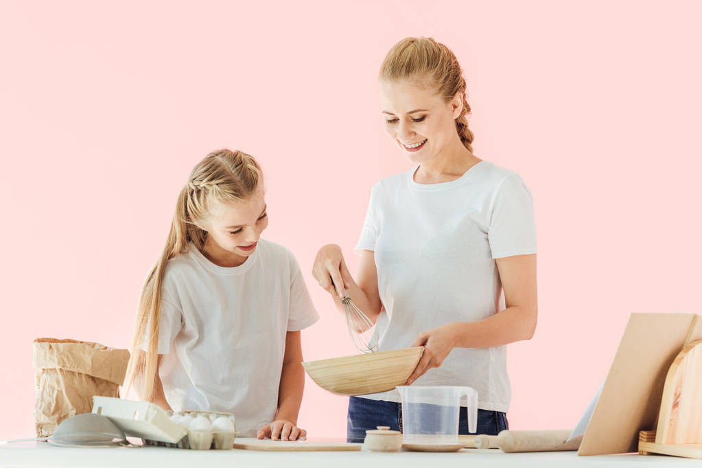 glückliche Mutter und Tochter in weißen T-Shirts beim gemeinsamen Kochen auf rosa Schale schauend - Foto, Bild