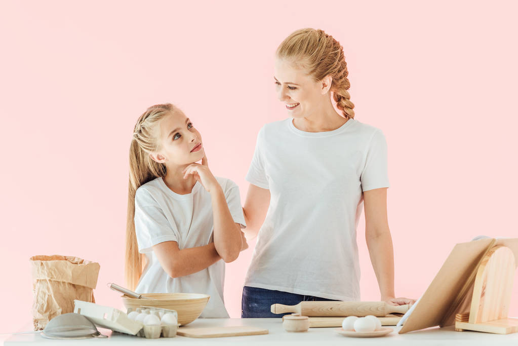 feliz madre e hija en camisetas blancas divirtiéndose juntas mientras cocinan aisladas en rosa
 - Foto, Imagen