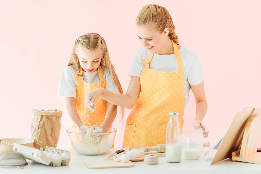 Улыбающаяся молодая мать и очаровательная маленькая дочь в желтых фартуках, вместе готовящая тесто, изолированное на розовом
 - Фото, изображение