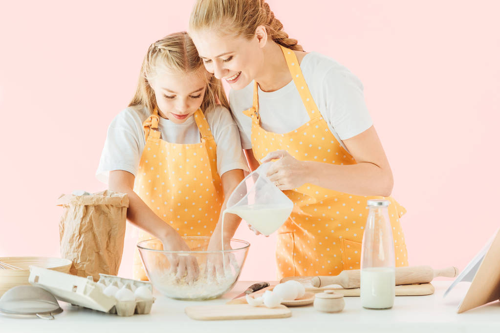 ευτυχισμένη μητέρα ρίχνει γάλα στην ζύμη, ενώ κόρη ζύμωμα απομονώνονται σε ροζ - Φωτογραφία, εικόνα
