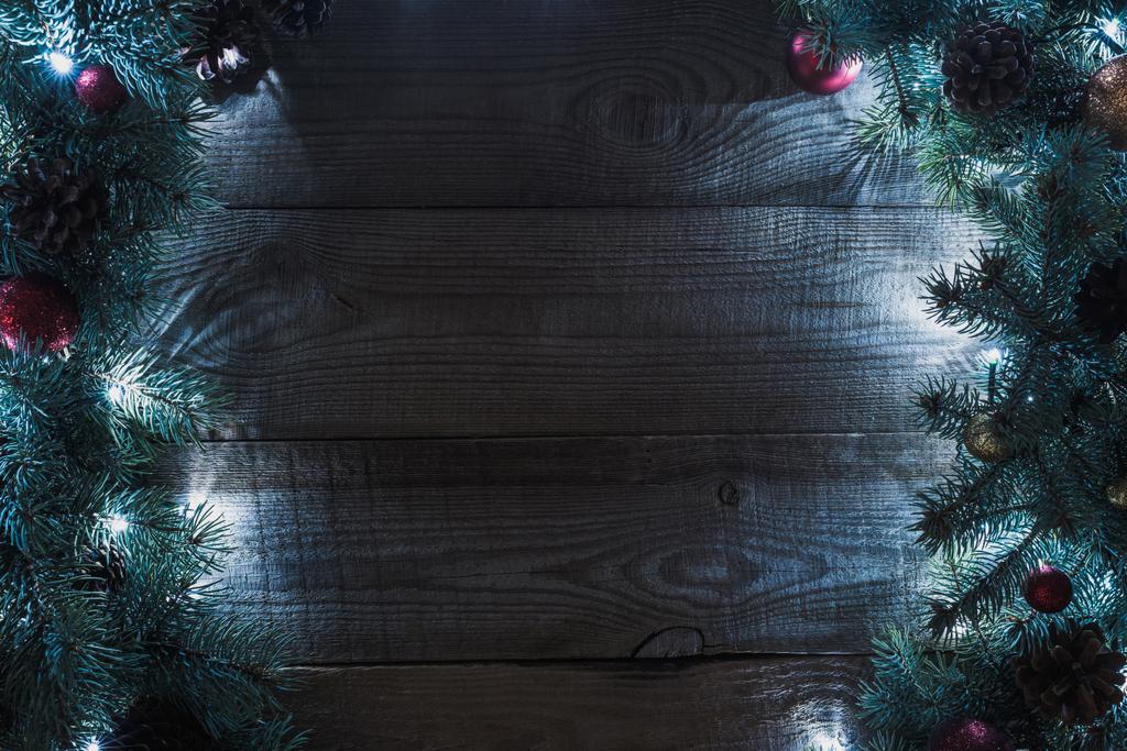 top view of christmas tree twigs with pine cones, baubles and illuminated garland on wooden background - Photo, Image