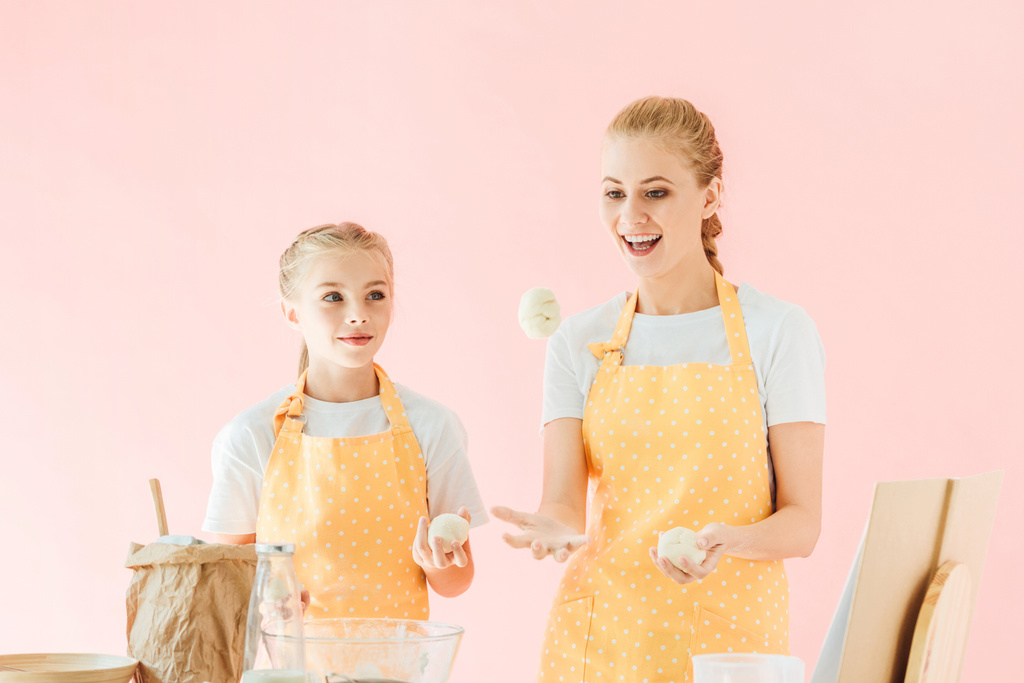 Lächelnde Mutter und Tochter jonglieren mit Teigstücken beim Kochen isoliert auf rosa - Foto, Bild