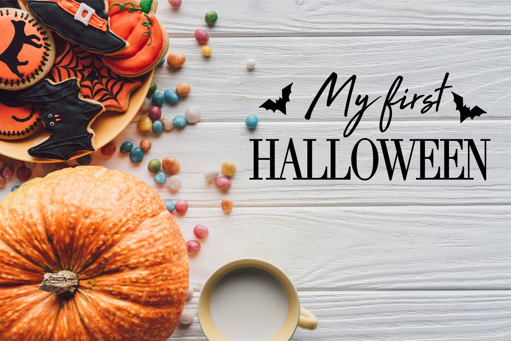 Verhoogd zicht op pompoen, bord met halloween koekjes, snoepjes en beker met melk op houten achtergrond met vleermuizen en "mijn eerste Halloween" belettering - Foto, afbeelding