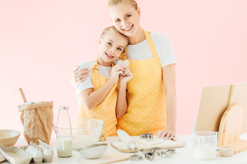 ευτυχισμένος νεαρή μητέρα και κόρη με ζύμη σε σχήμα καρδιάς βλέπουν κάμερα ενώ ετοιμάζει cookies που απομονώνονται σε ροζ - Φωτογραφία, εικόνα