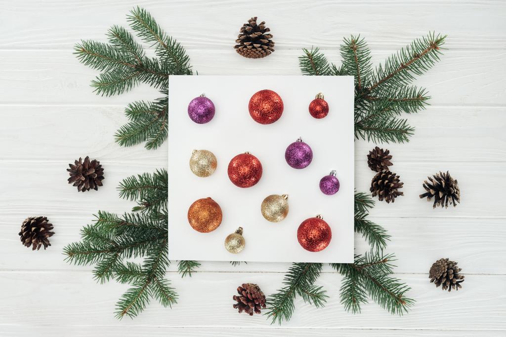 το Top view λαμπερά πολύχρωμες Χριστουγεννιάτικες μπάλες από λευκή κάρτα, έλατο κλαδιά και κουκουνάρια σε ξύλινη επιφάνεια - Φωτογραφία, εικόνα