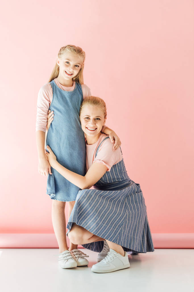 Lächelnde Mutter hockt in der Hocke und umarmt ihre Tochter auf rosa und blickt in die Kamera - Foto, Bild