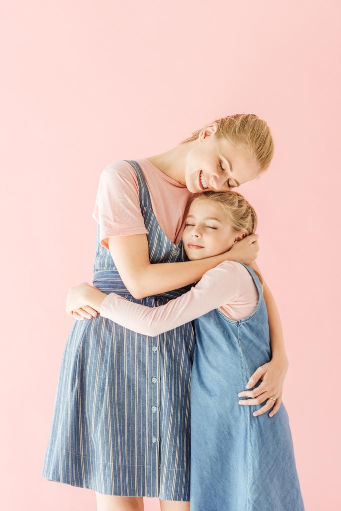 Gelukkig jonge moeder en dochter in het blauw jurken omarmen geïsoleerd op roze - Foto, afbeelding