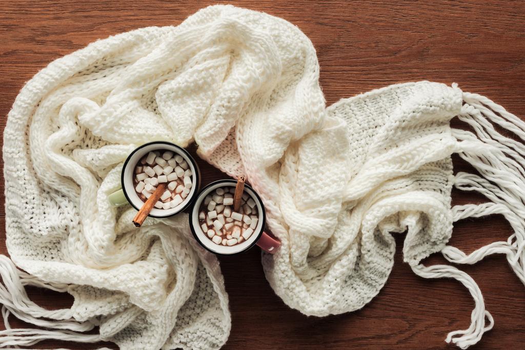 flat lay with mugs with hot chocolate, marshmallows and cinnamon sticks, white knitted scarf on wooden surface - Photo, Image