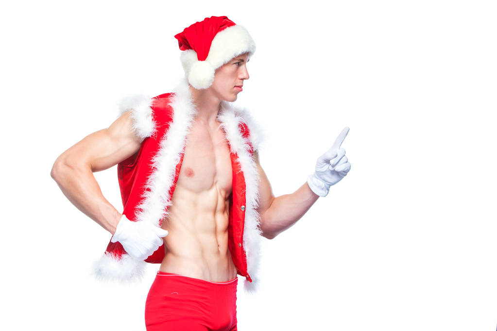 Sexy Santa Claus señalando en blanco signo en blanco. Un joven musculoso con sombrero de Santa Claus demuestra sus músculos. Aislado sobre fondo blanco
. - Foto, Imagen