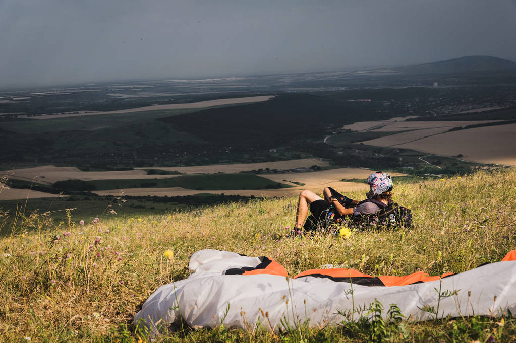 Un parapente professionnel en équipement complet et un casque repose et repose sur l'herbe haute dans les montagnes en regardant les nuages
 - Photo, image