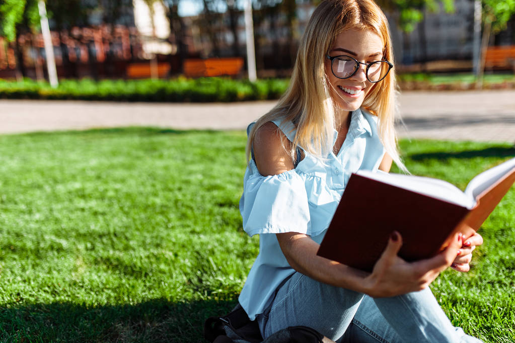 Portrét veselý student dívka sedí venku na zelené trávě, nosí brýle, drží knihu a čtení - Fotografie, Obrázek