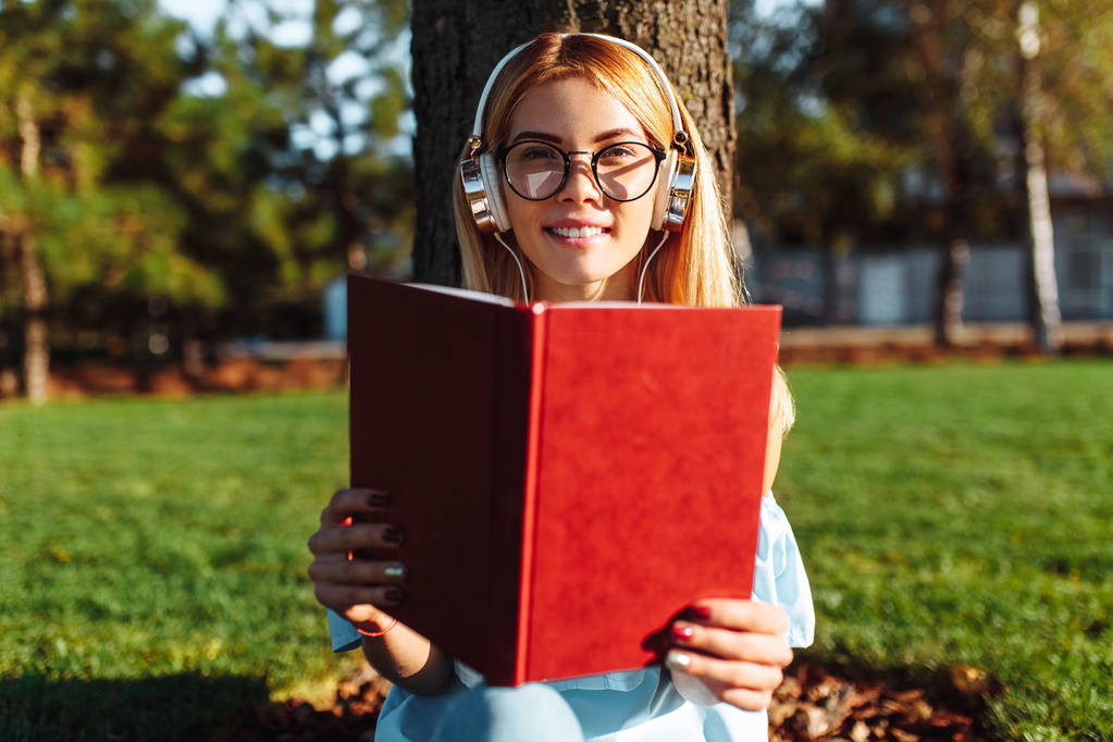 Portrét veselá krásnou coed dívky, poslechu hudby ve sluchátkách, sedí venku na zelené trávě, nosí brýle, drží knihu a čtení - Fotografie, Obrázek