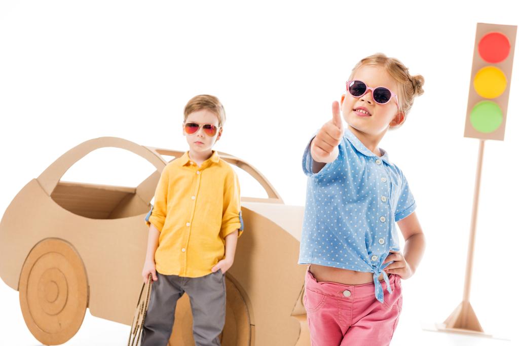 αξιολάτρευτο παιδί κομψό σε γυαλιά ηλίου που δείχνει τον αντίχειρα επάνω ενώ στέκεται κοντά στο αυτοκίνητο από χαρτόνι και φώτα κυκλοφορίας, σε λευκό αγόρι  - Φωτογραφία, εικόνα